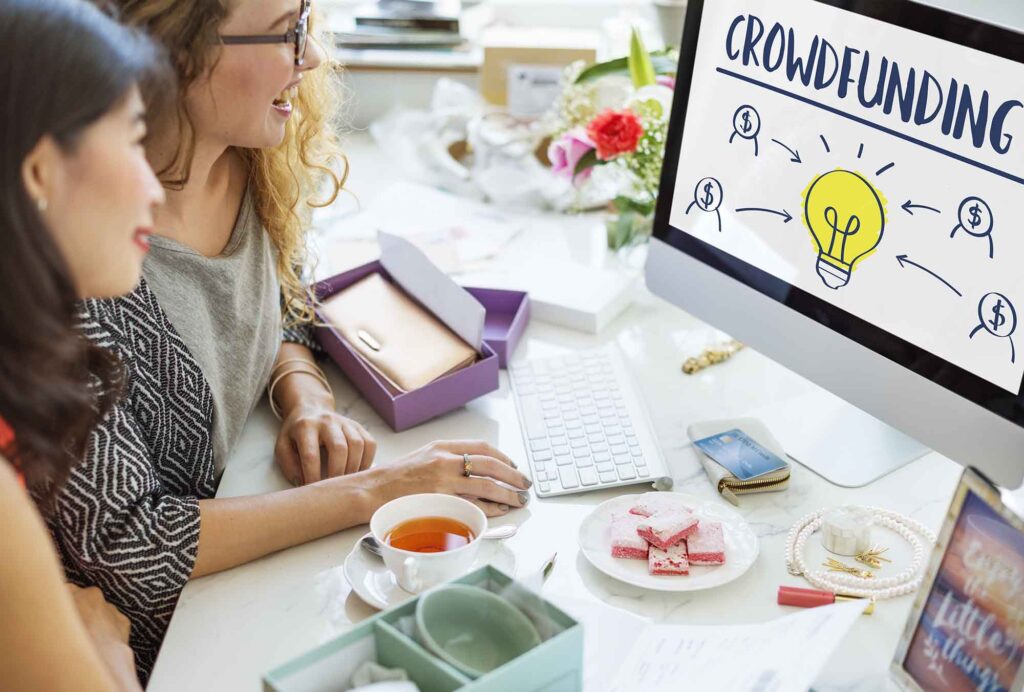 Crowdfunding Money Business Bulb Graphic Concept | AD-IOS Web Development Two women review a crowdfunding concept on a computer monitor during a bright, casual meeting with snacks nearby
