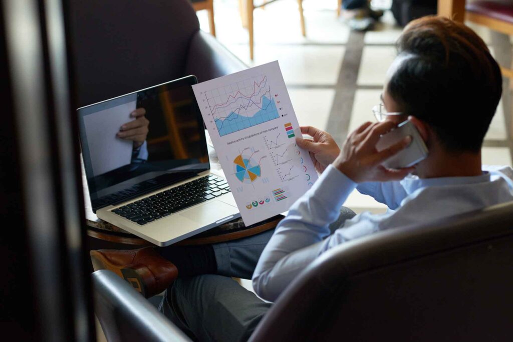 Working with financial document | AD-IOS Web Development Person sits at a desk with a laptop, studying a printed data report filled with colorful charts while talking on a phone.