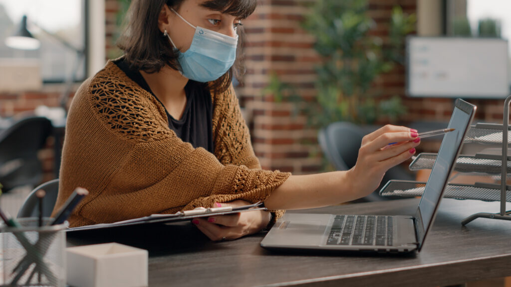 Close up of woman using papers and laptop to work | AD-IOS Web Development Person wearing a blue mask at a desk, using a tablet and laptop with a pen nearby, in an office setting.