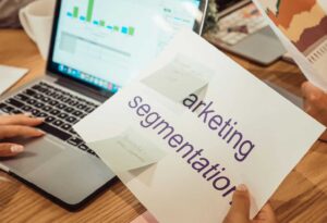Digital Marketing-img2 | AD-IOS Web Development Person holds a brochure titled'marketing segmentation' over a laptop showing charts on the screen, with sticky notes nearby on a desk.