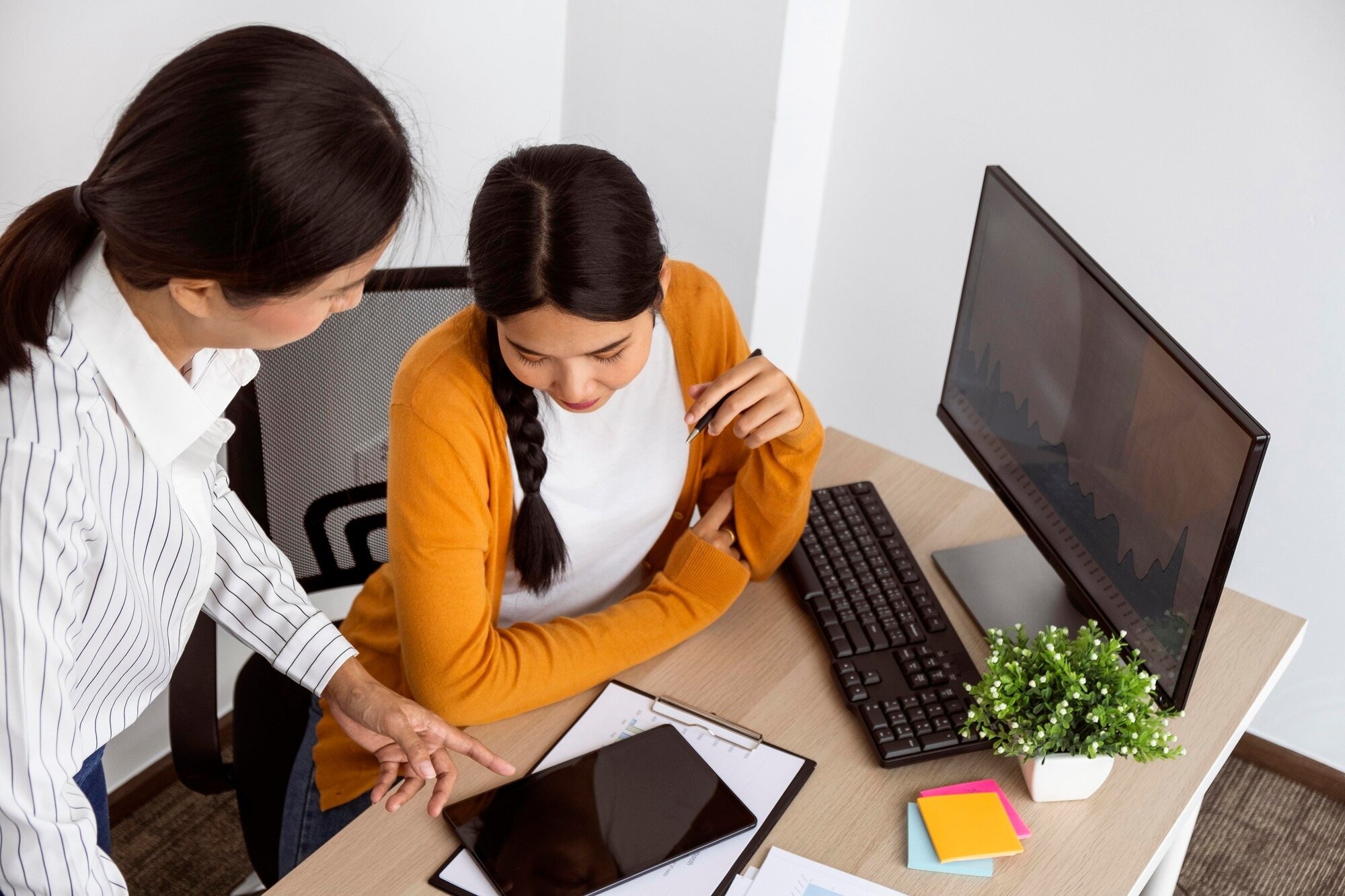 Employee Onboarding Software 2 | AD-IOS Web Development Two women collaborate at a desk: one points at a tablet while the other looks on, with a monitor, keyboard, papers, and colorful sticky notes nearby.