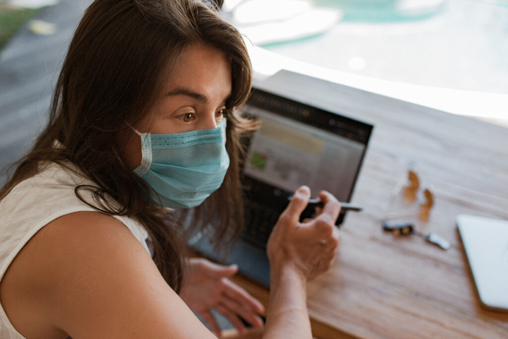 girl working on laptop in a mask | AD-IOS Web Development Woman wearing a blue surgical mask, looking at a smartphone while seated at a wooden table.